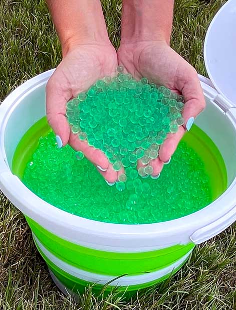 Hands holding green water beads over a bucket filled with them